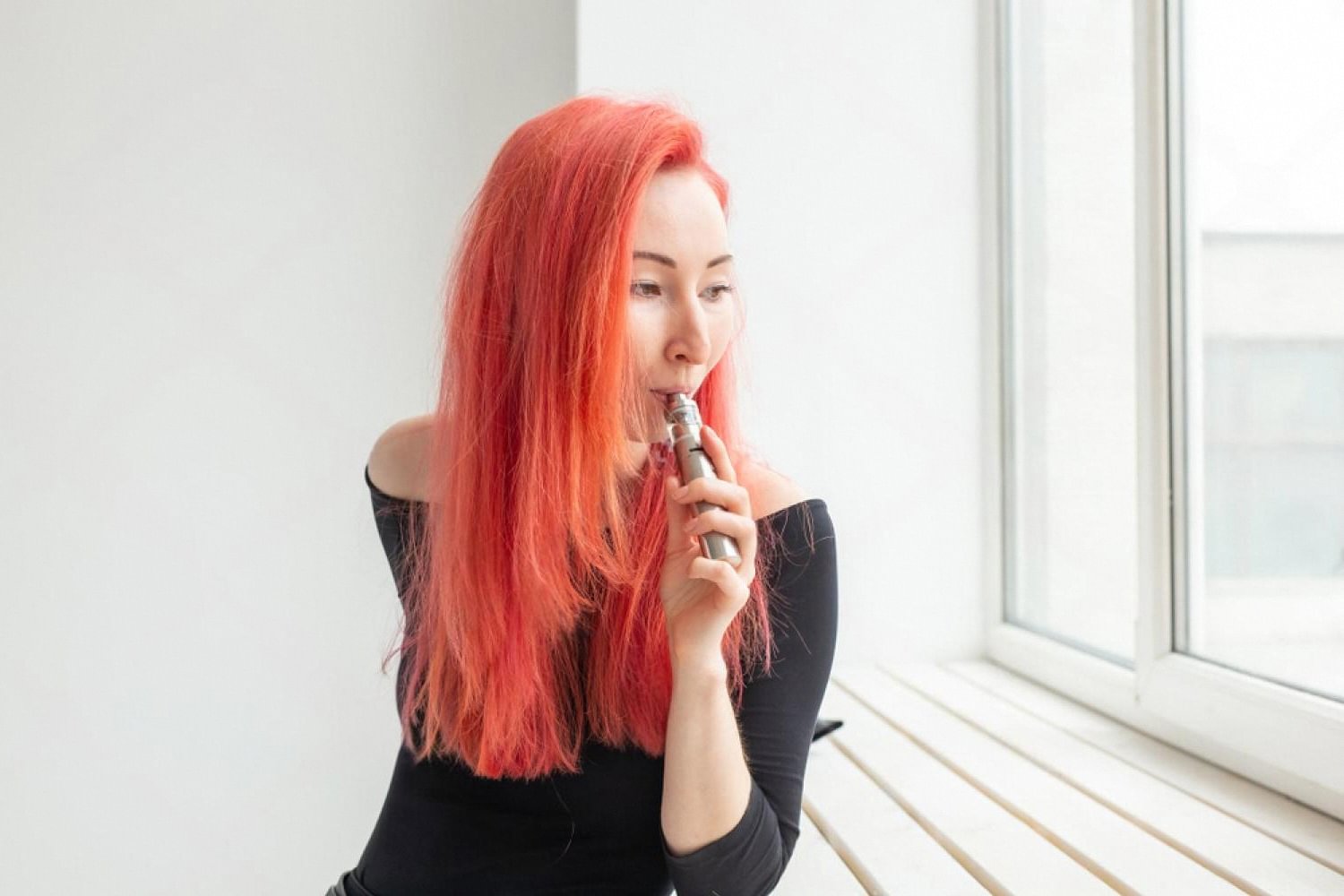 Woman with red hair using a vape device near a bright window